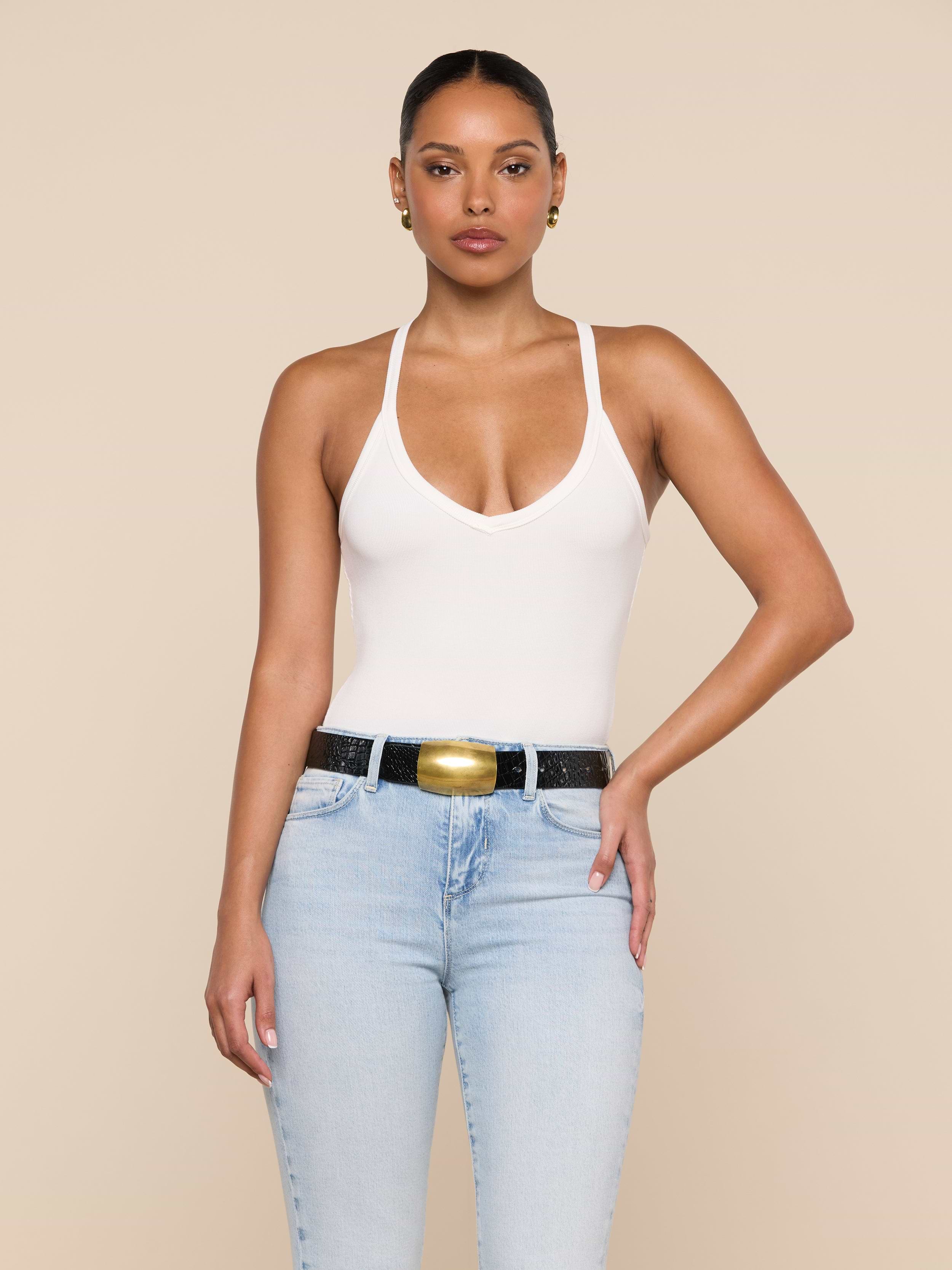 A woman stands against a beige background, wearing the Wrenna Racerback Bodysuit in white rib fabric, light blue jeans, a black belt with a gold buckle, and gold earrings. Her hair is pulled back and one hand rests on her hip.