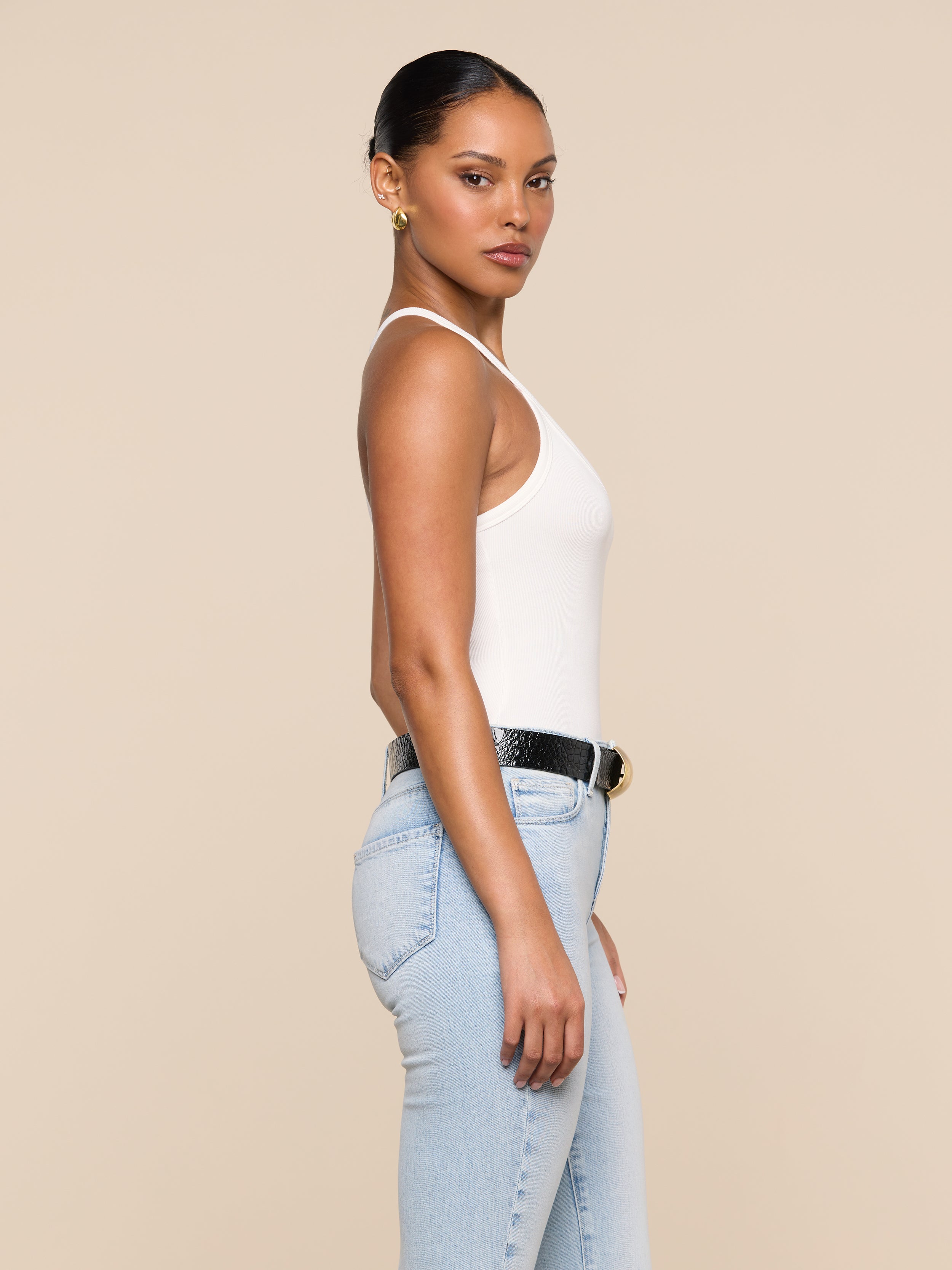 A woman stands in profile against a beige background, wearing the Wrenna Racerback Bodysuit in white rib fabric, light blue jeans, a black belt with a gold buckle, and gold hoop earrings. Her hair is pulled back and she has a neutral expression.