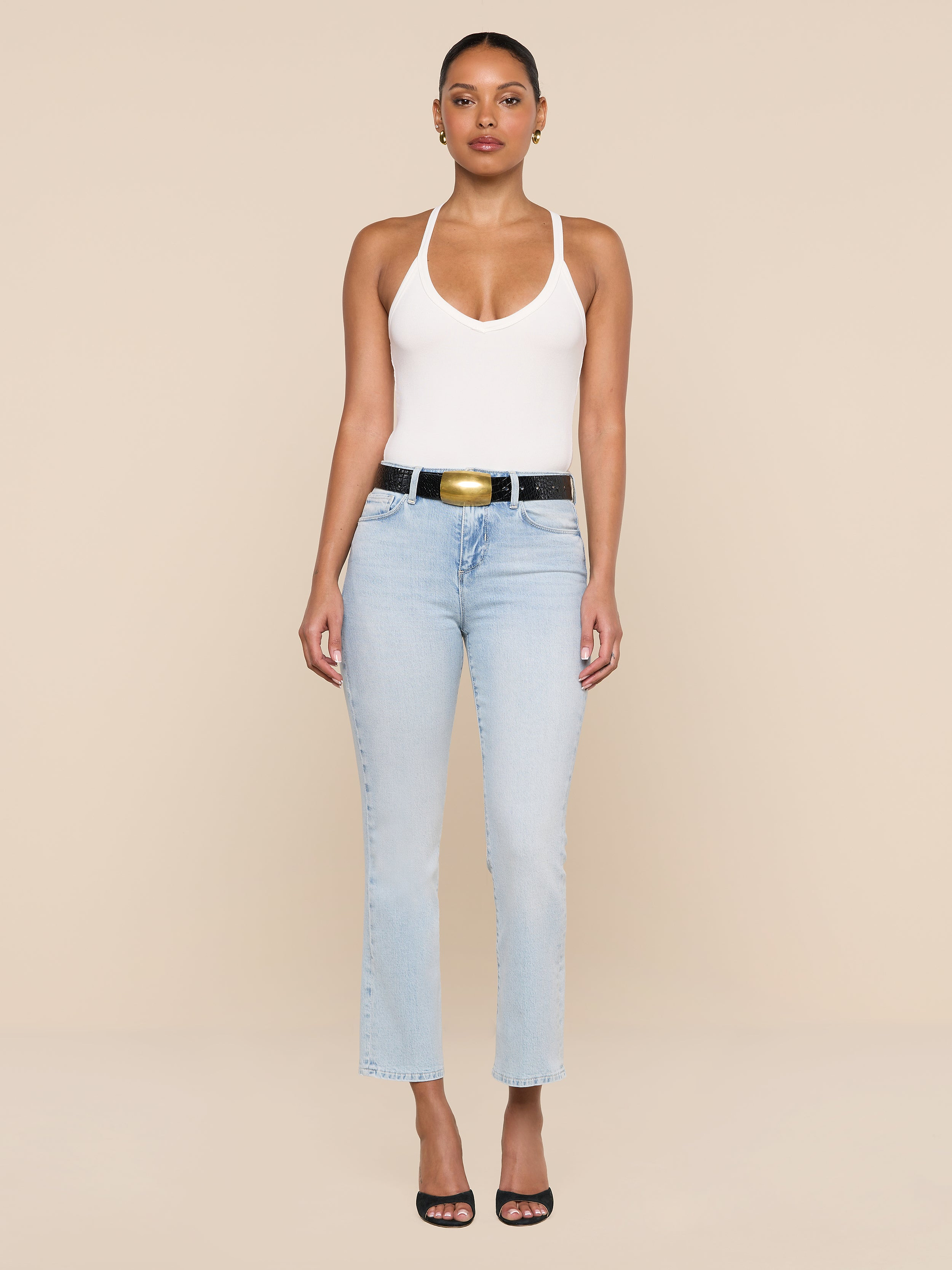 A woman stands against a beige background, wearing the Wrenna Racerback Bodysuit in white rib fabric, light blue high-waisted jeans, a black belt with a gold buckle, and black open-toe heels. She faces forward with arms at her sides.