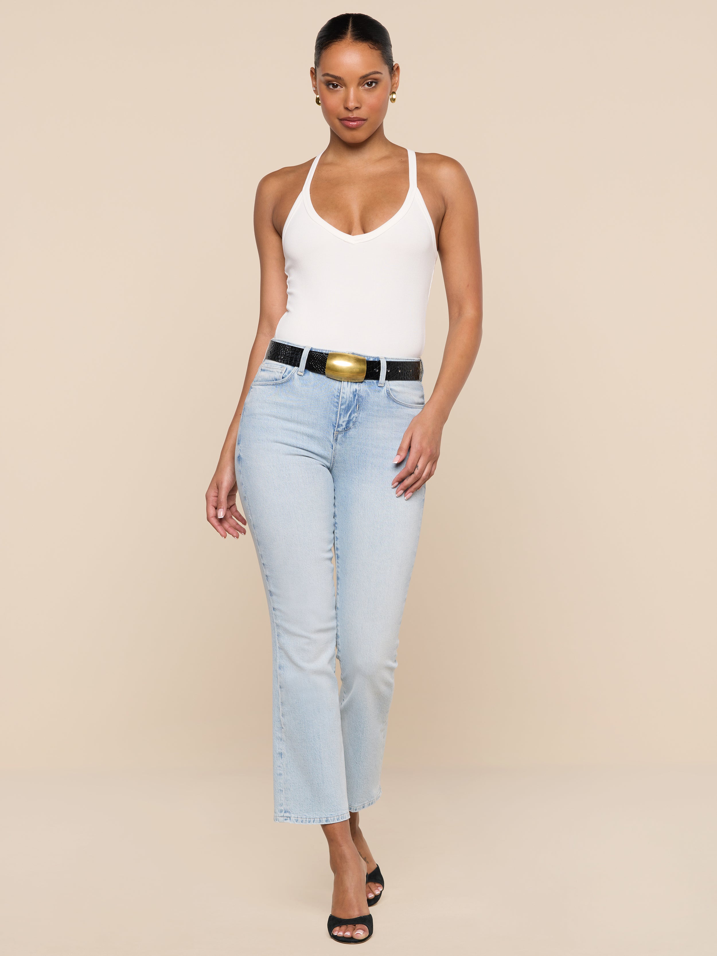 A woman poses against a beige background wearing the Wrenna Racerback Bodysuit in white, paired with light blue high-waisted jeans, a black belt, and black heeled sandals.
