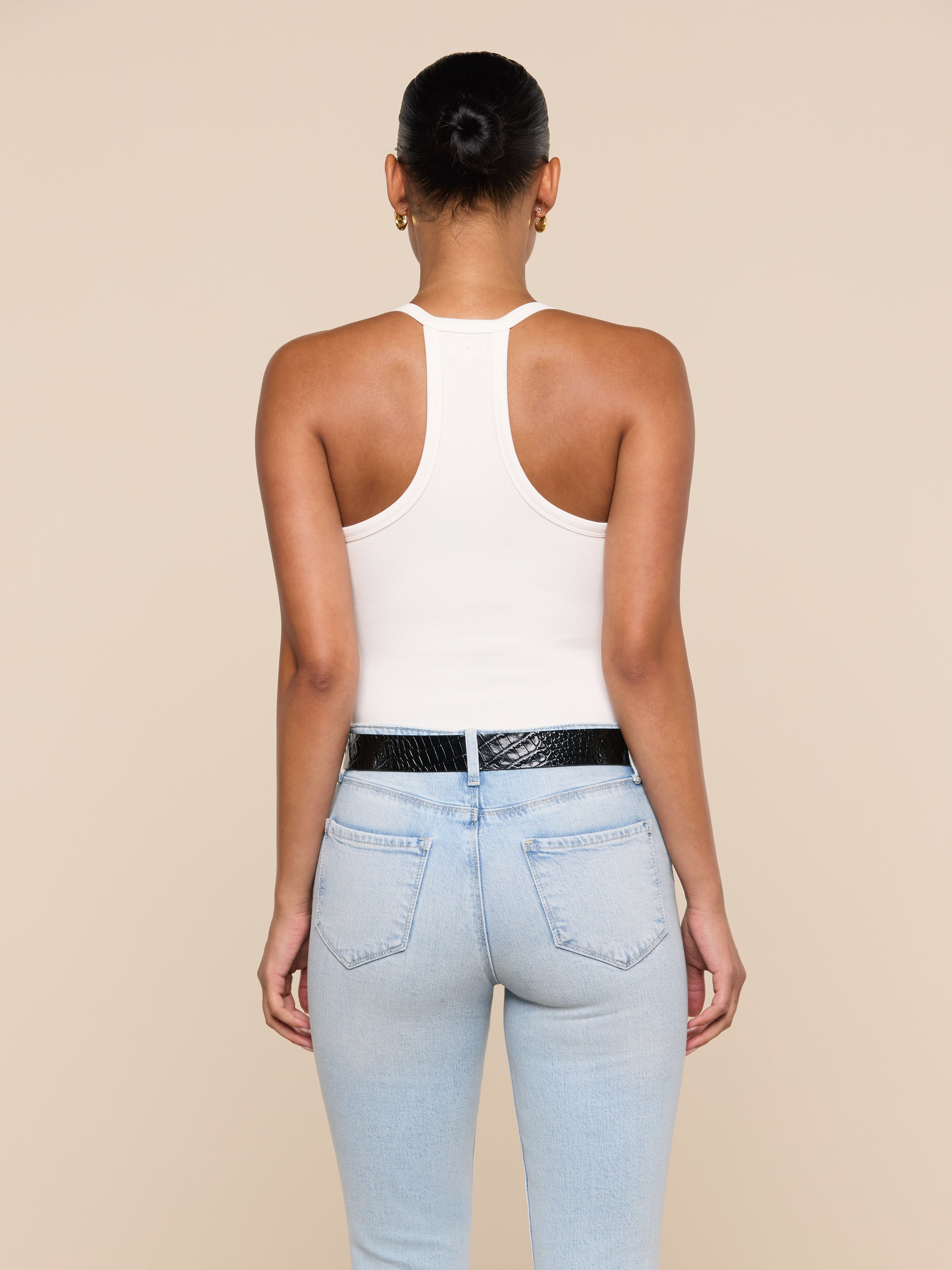 A person is shown from the back wearing the Wrenna Racerback Bodysuit in white, paired with light blue jeans, a black belt, and small hoop earrings. Their hair is in a neat bun against a plain beige background.