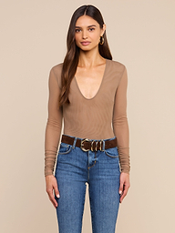 A woman with long brown hair is standing against a plain light background, wearing the Thalassa High-Leg Bodysuit featuring sexy mesh and sheer nude long sleeves, paired with blue jeans, a wide brown belt with large gold buckles, and gold hoop earrings.