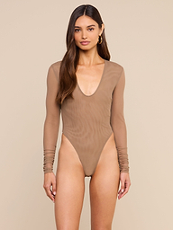 A woman models the Thalassa High-Leg Bodysuit, featuring sheer mesh fabric, long sleeves, and a deep scoop neckline. She stands against a plain light background, her long brown hair cascading over her shoulders as she looks at the camera.