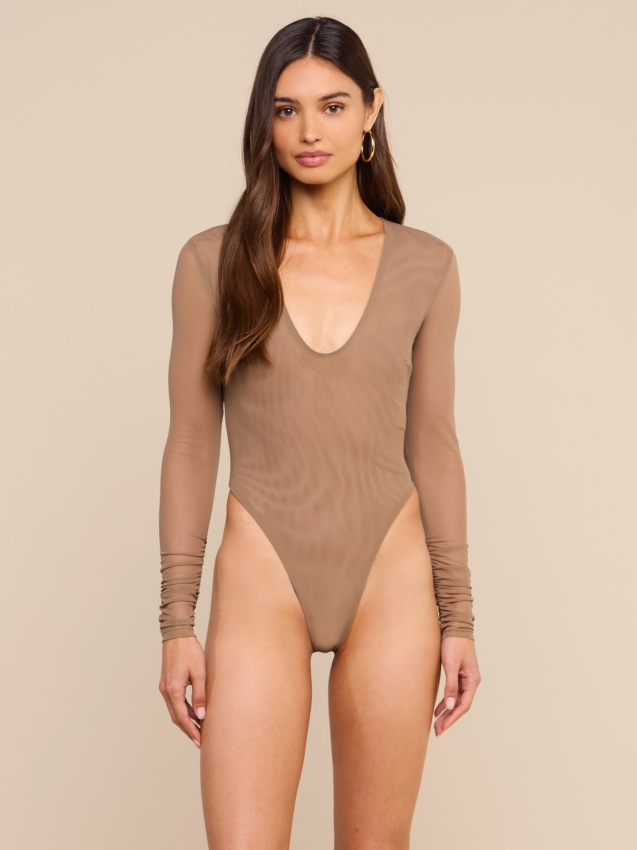 A woman models the Thalassa High-Leg Bodysuit, featuring sheer mesh fabric, long sleeves, and a deep scoop neckline. She stands against a plain light background, her long brown hair cascading over her shoulders as she looks at the camera.