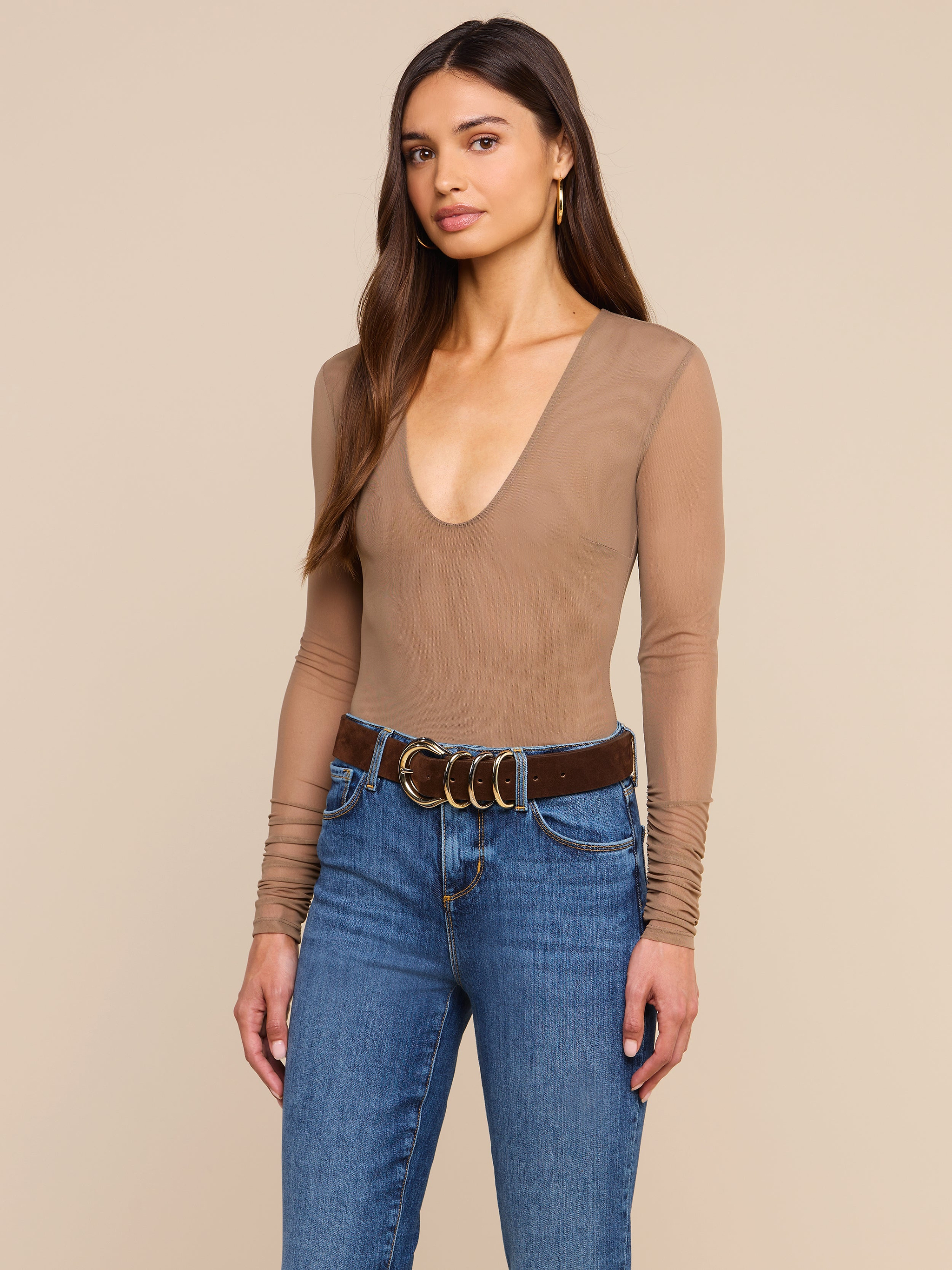 A woman with long brown hair wears the Thalassa High-Leg Bodysuit paired with blue jeans and a brown belt, standing against a plain light background.