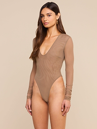 A woman stands against a plain background wearing the Thalassa High-Leg Bodysuit—a fitted, long-sleeve mesh piece with a deep neckline in sheer beige. She has long brown hair and a neutral expression, embodying effortless French style.
