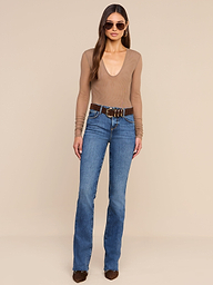 A woman stands against a plain background, wearing sunglasses, blue jeans with a brown belt, brown shoes, and the Thalassa High-Leg Bodysuit in tan.