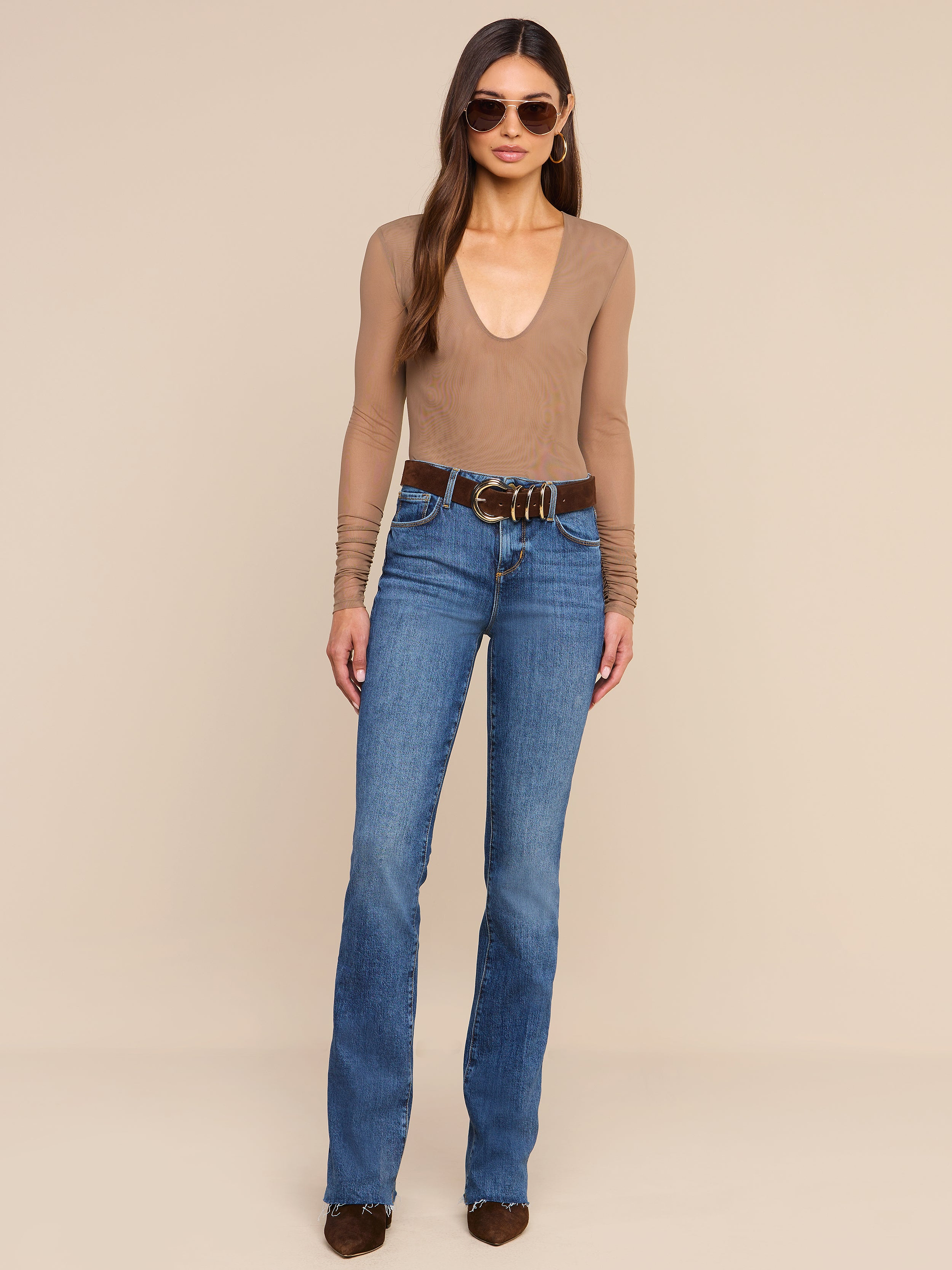 A woman stands against a plain background, wearing sunglasses, blue jeans with a brown belt, brown shoes, and the Thalassa High-Leg Bodysuit in tan.