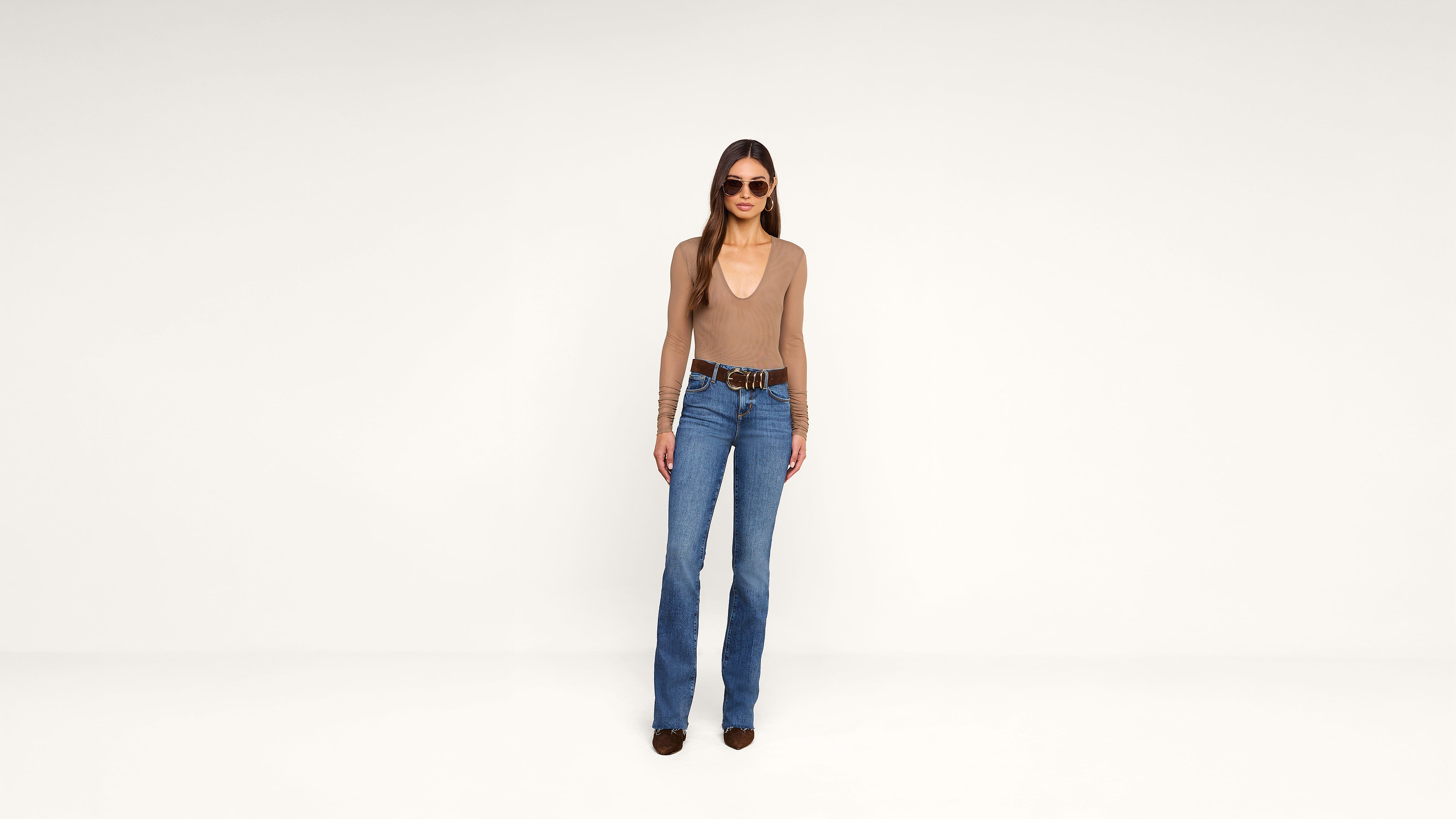 Woman standing against a white background wearing sunglasses, a tan long-sleeve top, blue jeans, a belt, and dark shoes.