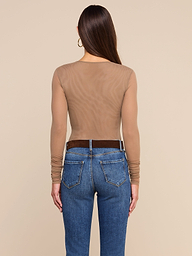 A woman stands facing away, wearing the Thalassa High-Leg Bodysuit—a sheer, long-sleeve mesh style—paired with blue jeans and a dark belt. Her long, straight hair is complemented by large hoop earrings. The background is plain and light-colored.