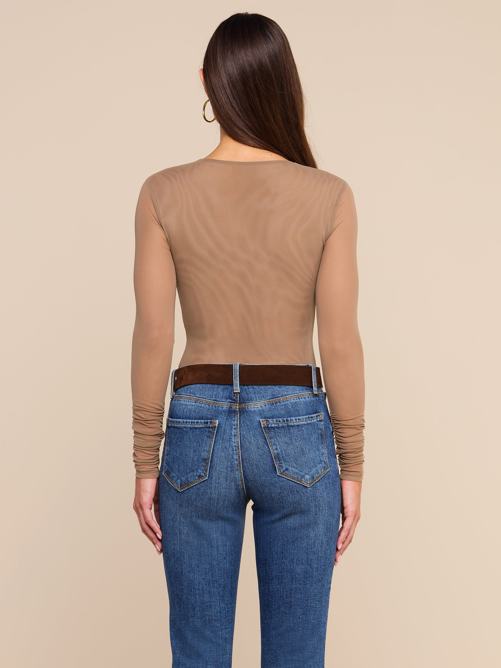 A woman stands facing away, wearing the Thalassa High-Leg Bodysuit—a sheer, long-sleeve mesh style—paired with blue jeans and a dark belt. Her long, straight hair is complemented by large hoop earrings. The background is plain and light-colored.