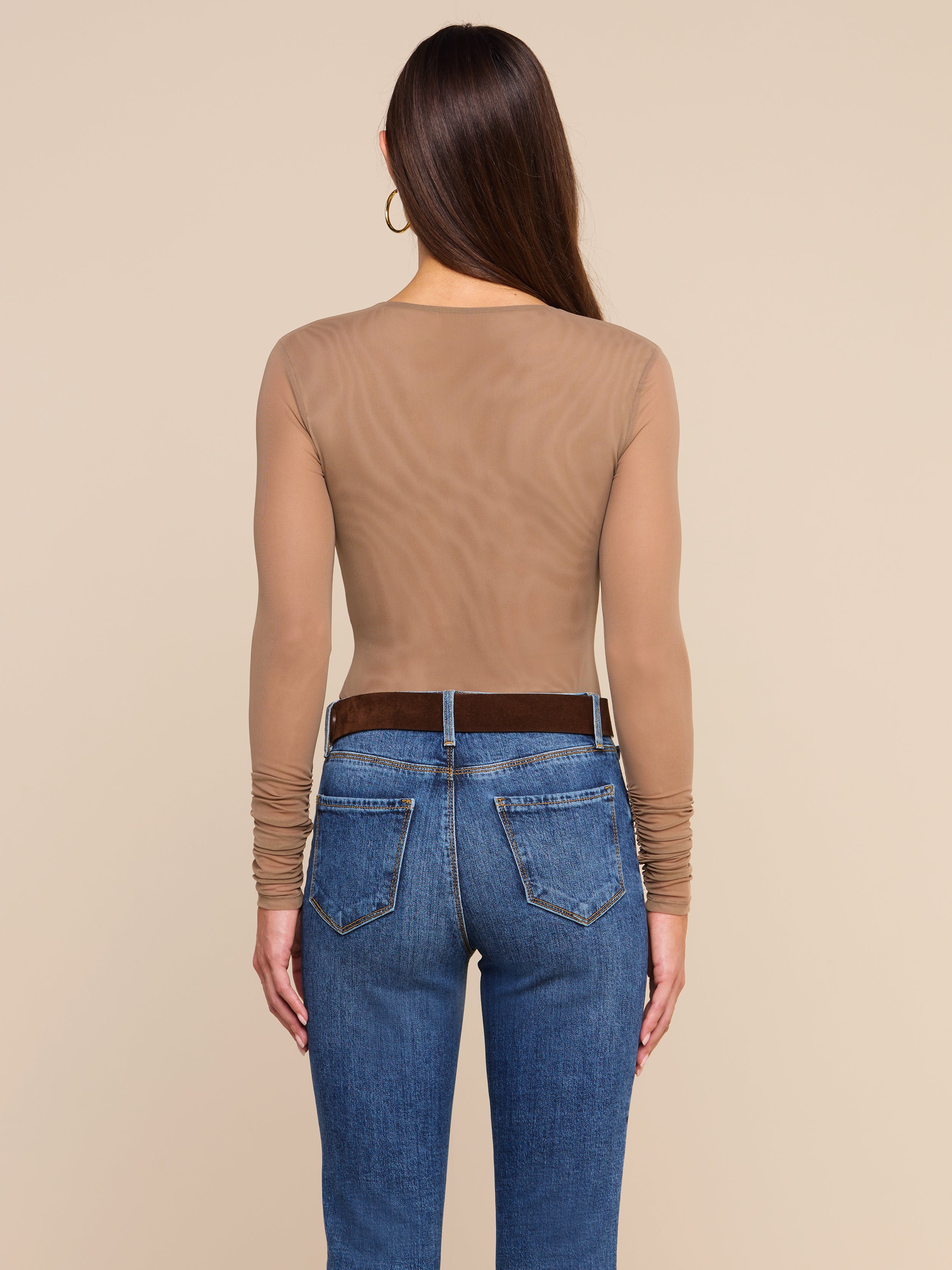 A woman stands facing away, wearing the Thalassa High-Leg Bodysuit—a sheer, long-sleeve mesh style—paired with blue jeans and a dark belt. Her long, straight hair is complemented by large hoop earrings. The background is plain and light-colored.
