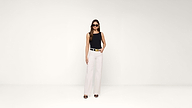 Woman wearing sunglasses, a sleeveless black top, white high-waisted pants, and black heels stands confidently against a plain white background with one hand in her pocket.