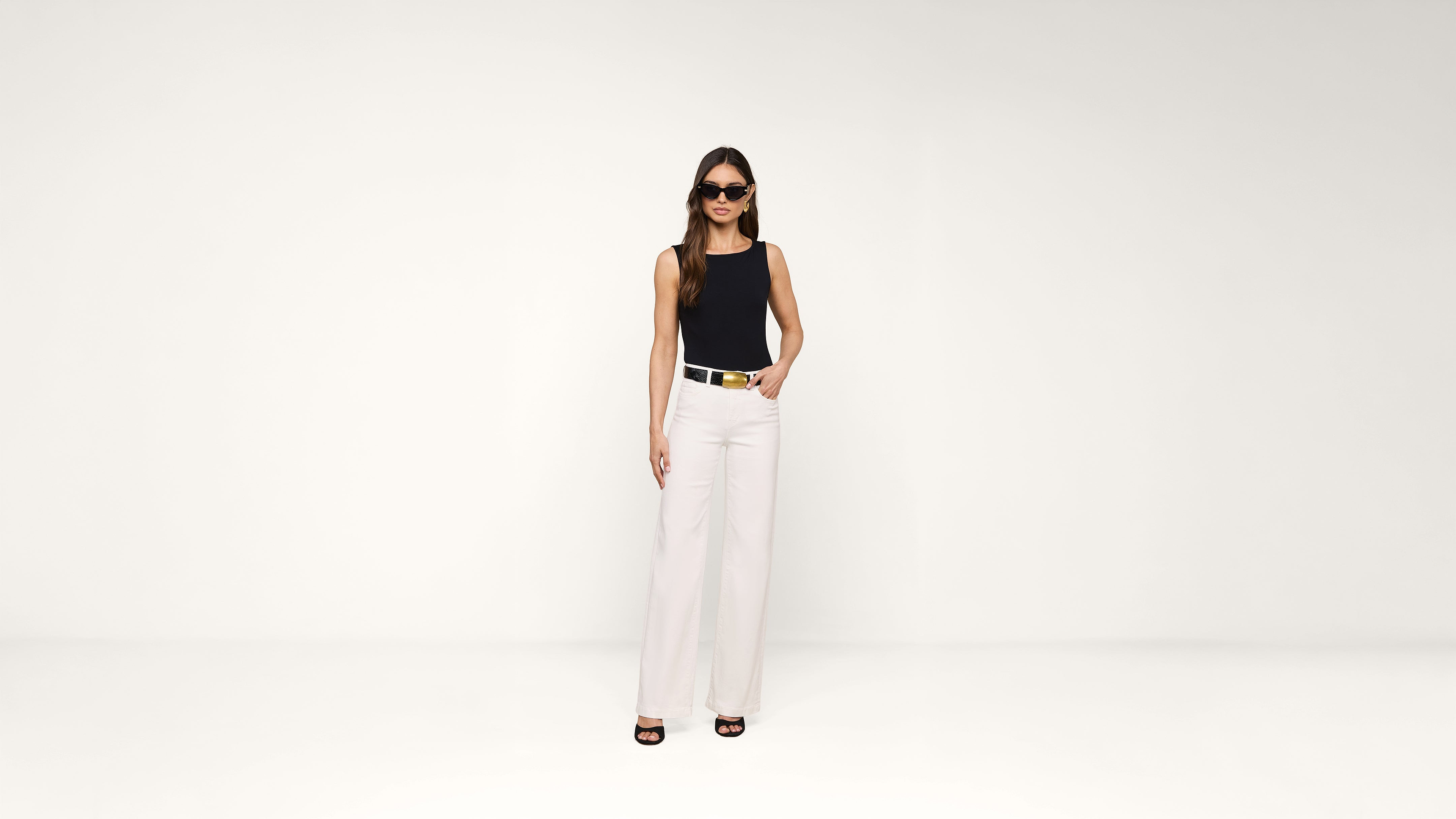 Woman wearing sunglasses, a sleeveless black top, white high-waisted pants, and black heels stands confidently against a plain white background with one hand in her pocket.