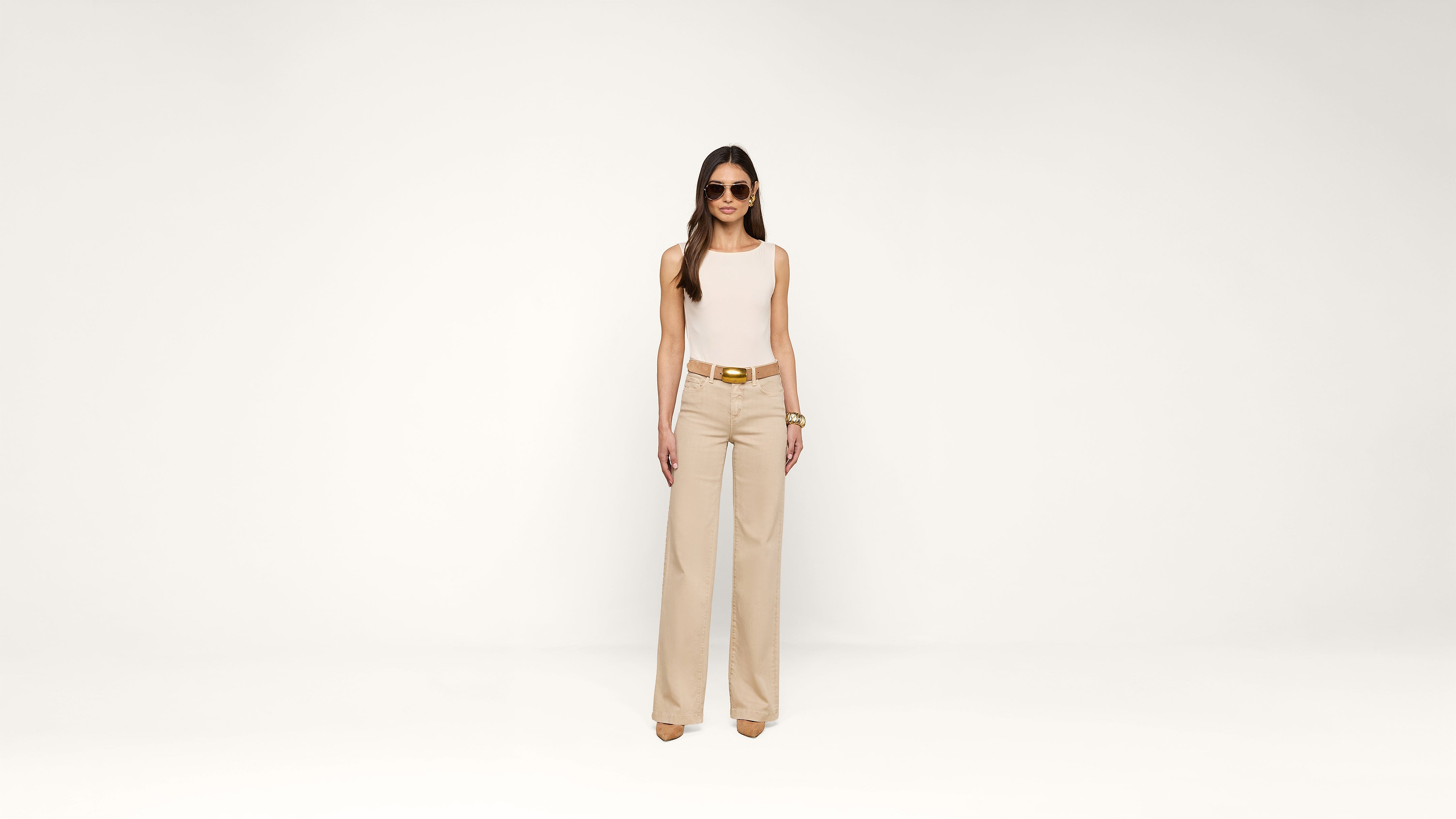 A woman wearing sunglasses, a sleeveless white top, beige wide-leg pants, and heels stands confidently against a plain white background.