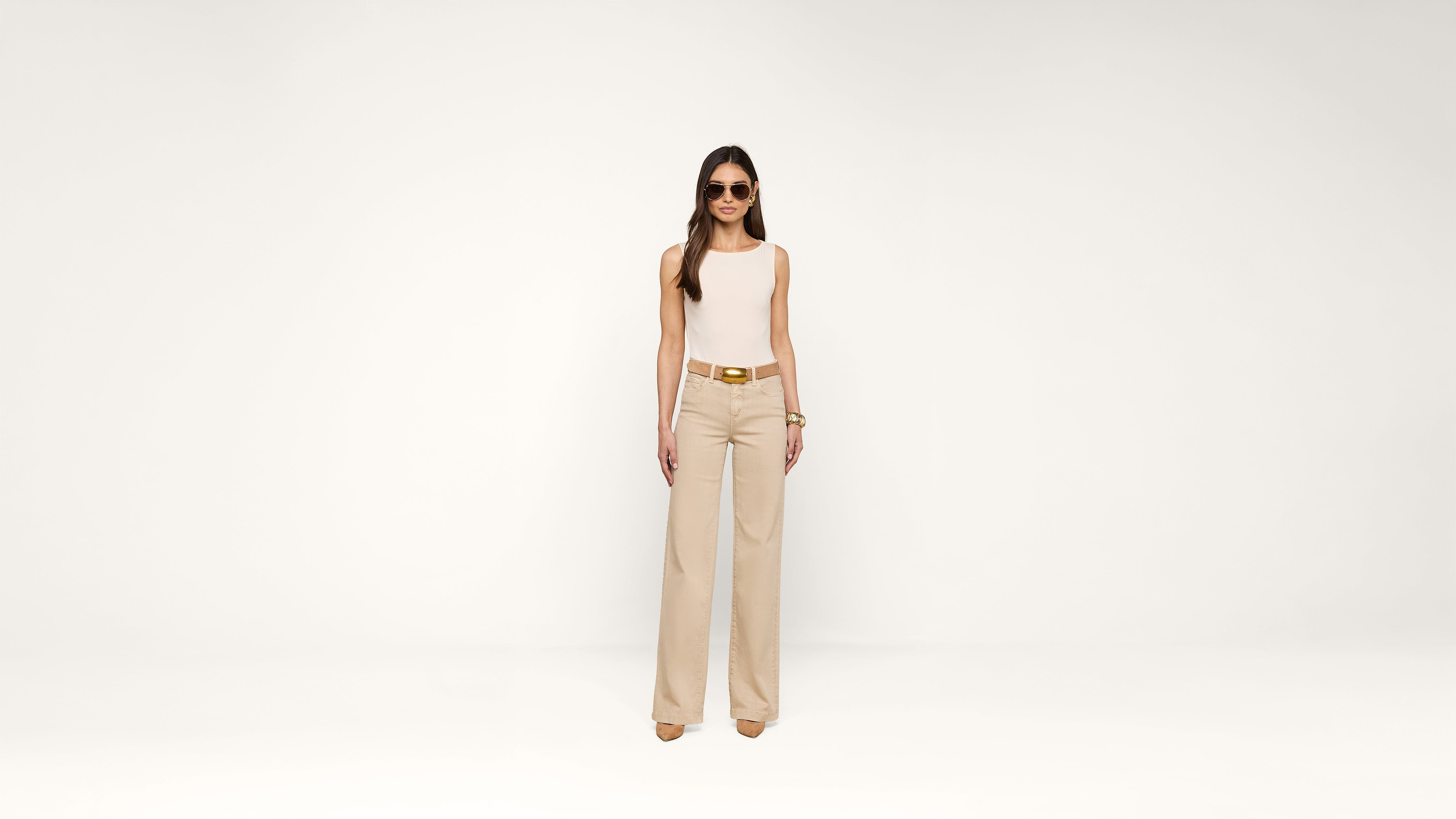 A woman wearing sunglasses, a sleeveless white top, beige wide-leg pants, and heels stands confidently against a plain white background.