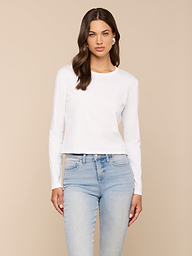 A woman with long brown hair wears the Rojene Cotton Tee, a relaxed fit plain white long sleeve shirt, paired with light blue jeans and standing against a plain light background.