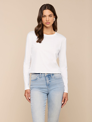 A woman with long brown hair wears the Rojene Cotton Tee in white with light blue jeans, standing against a beige background and looking at the camera with a neutral expression.