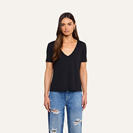 A woman with long brown hair, wearing a black Brianna Cotton V-Neck Tee in Supima cotton and ripped blue jeans, stands facing forward against a plain beige background.