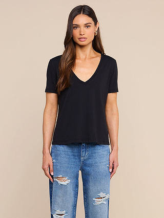 A woman with long brown hair, wearing a black Brianna Cotton V-Neck Tee in Supima cotton and ripped blue jeans, stands facing forward against a plain beige background.