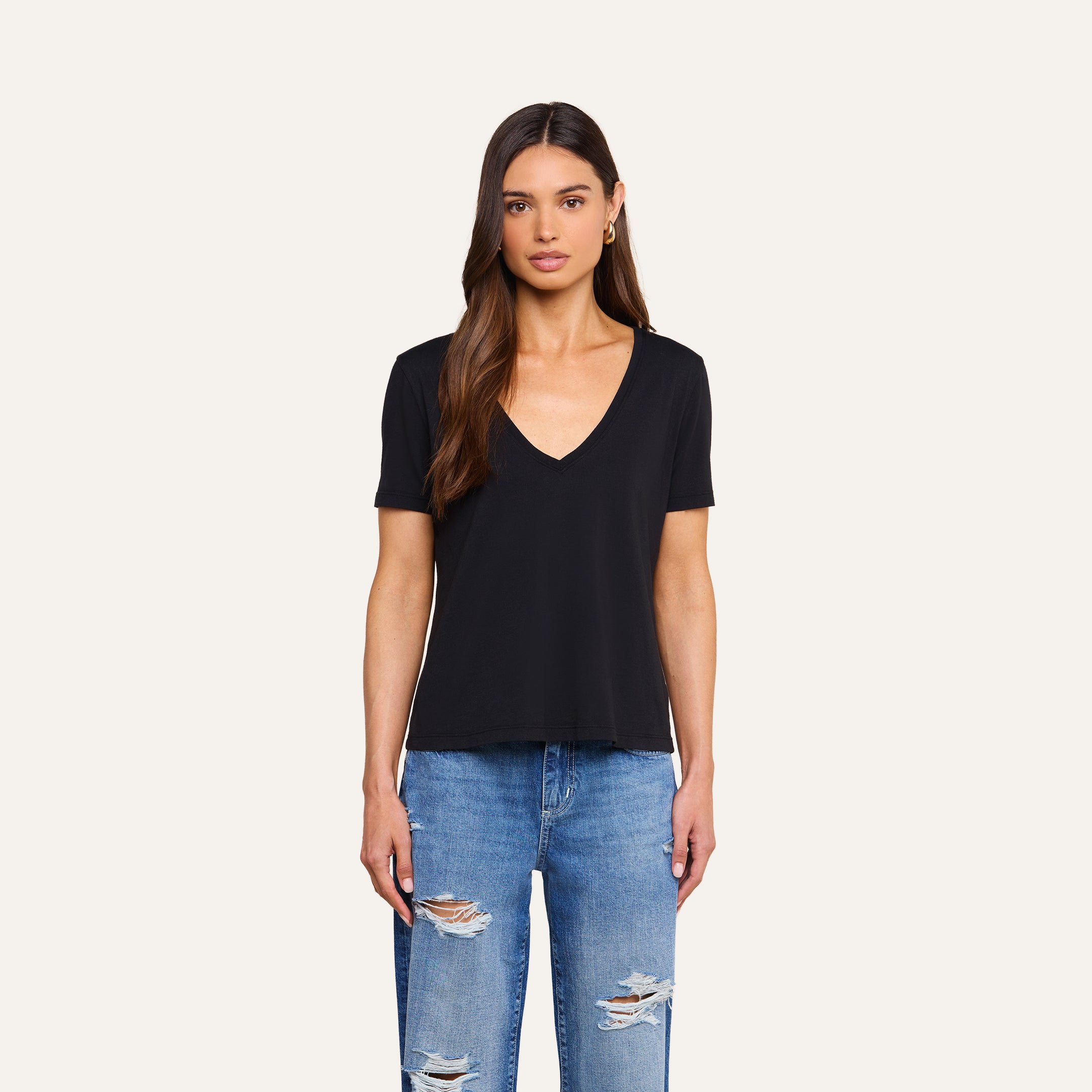 A woman with long brown hair, wearing a black Brianna Cotton V-Neck Tee in Supima cotton and ripped blue jeans, stands facing forward against a plain beige background.
