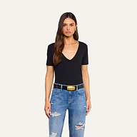 A woman with long brown hair wears a black Brianna Cotton V-Neck Tee made from soft Supima cotton, distressed blue jeans, a black belt with a gold buckle, and gold hoop earrings, standing against a plain beige background.