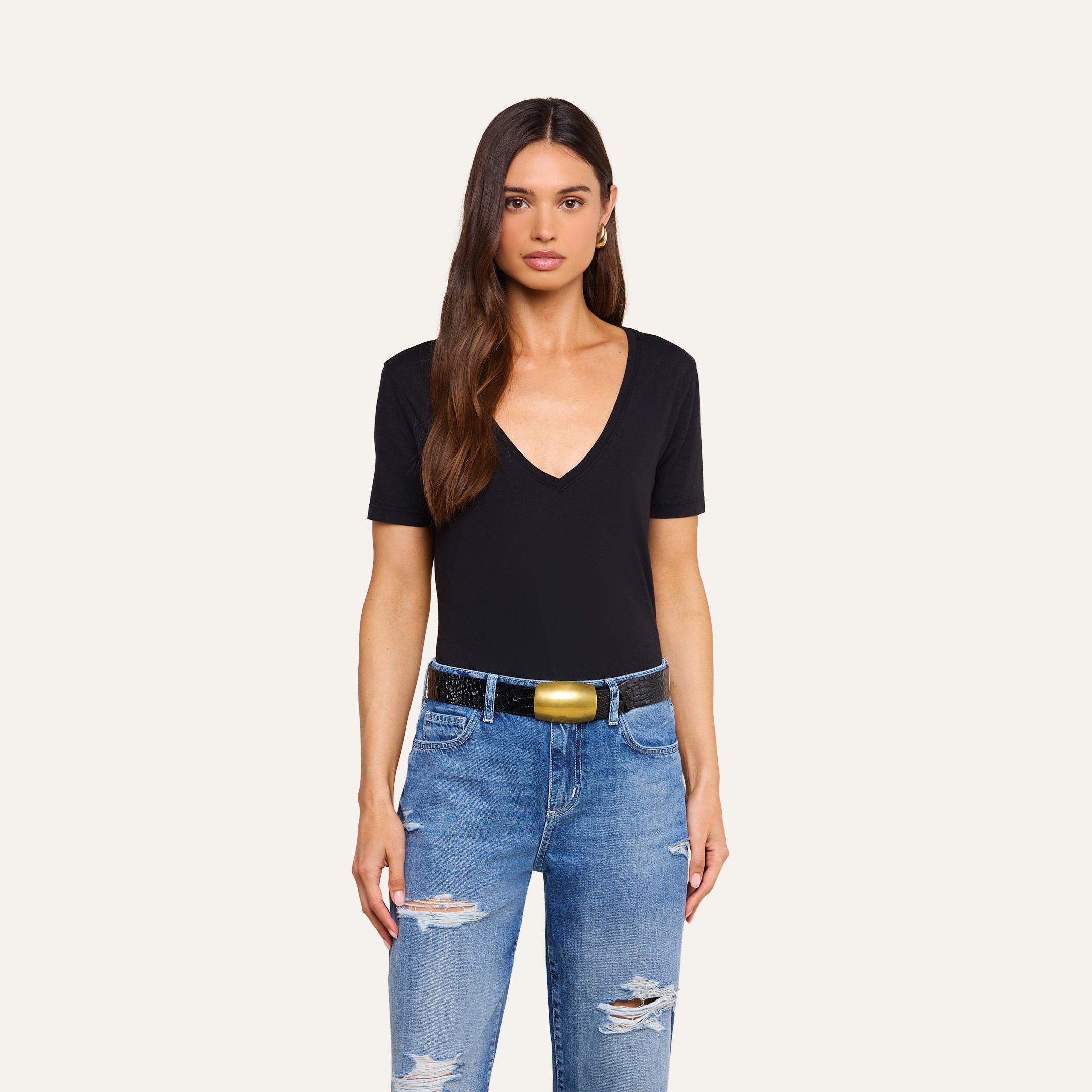 A woman with long brown hair wears a black Brianna Cotton V-Neck Tee made from soft Supima cotton, distressed blue jeans, a black belt with a gold buckle, and gold hoop earrings, standing against a plain beige background.
