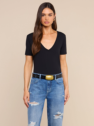 A woman with long brown hair wears a black Brianna Cotton V-Neck Tee made from soft Supima cotton, distressed blue jeans, a black belt with a gold buckle, and gold hoop earrings, standing against a plain beige background.