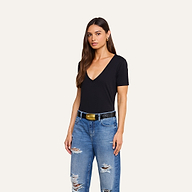 A woman with long brown hair is wearing a black Brianna Cotton V-Neck Tee, blue ripped jeans, and a black belt with a gold buckle, standing against a plain beige background.
