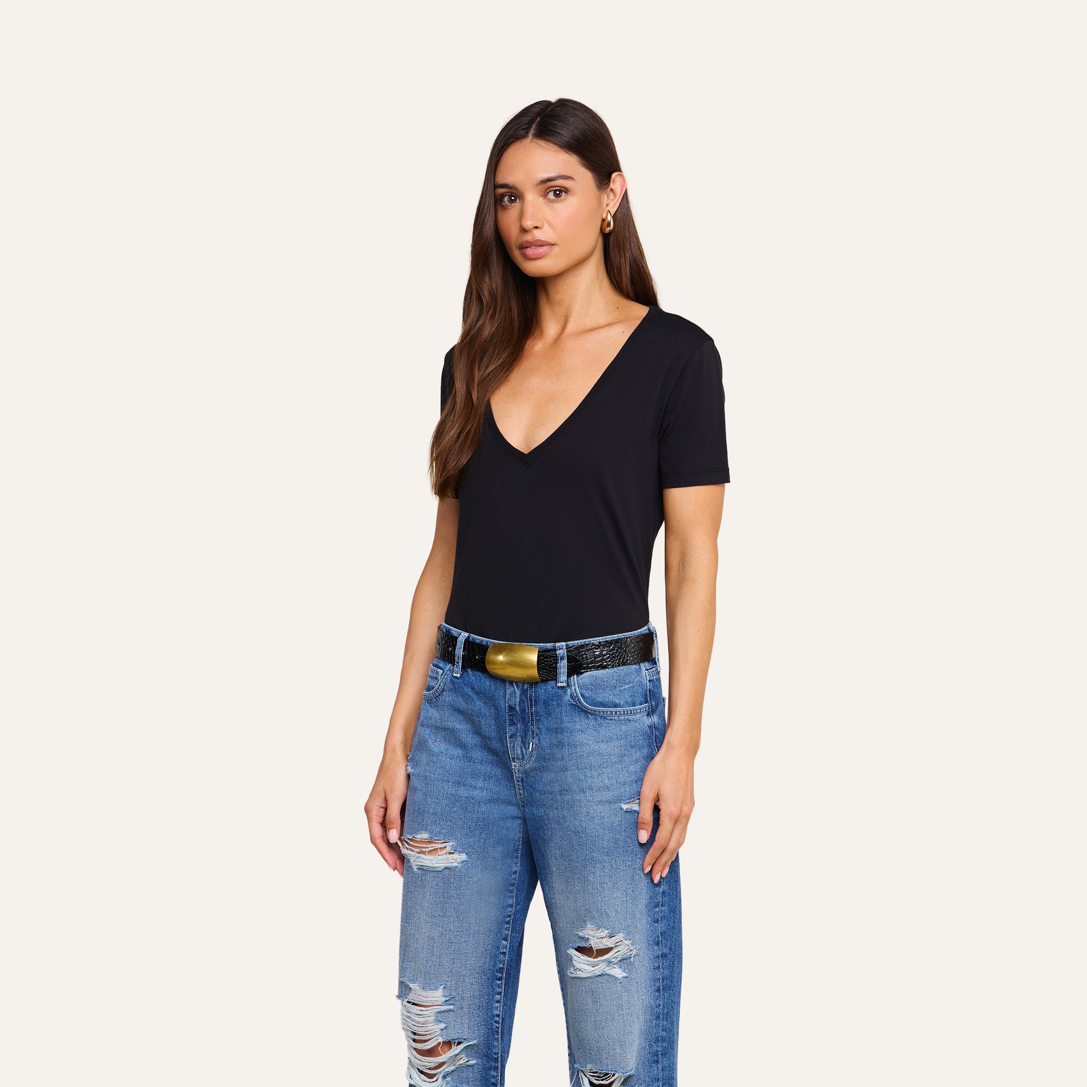 A woman with long brown hair is wearing a black Brianna Cotton V-Neck Tee, blue ripped jeans, and a black belt with a gold buckle, standing against a plain beige background.