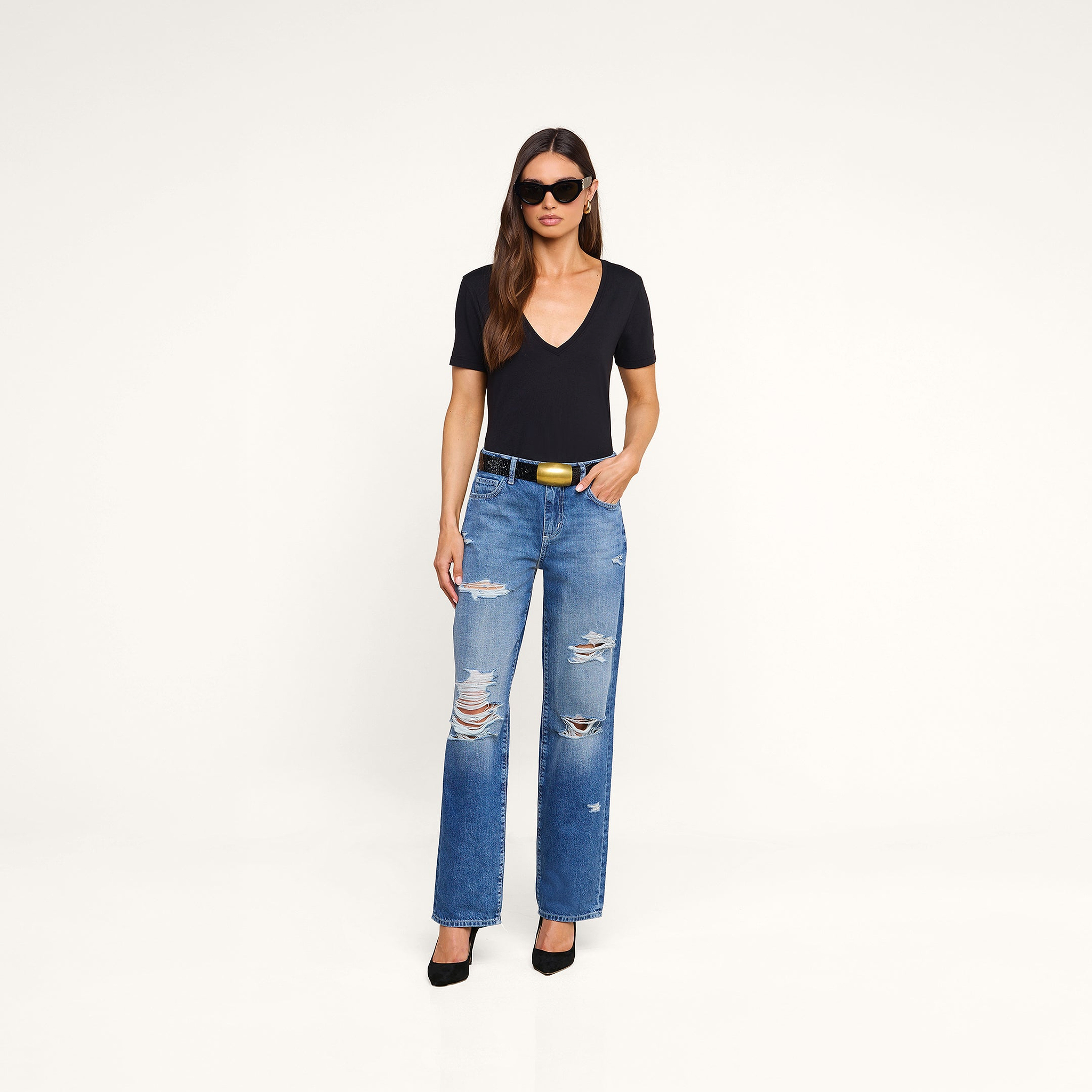 The woman is wearing the Brianna Cotton V-Neck Tee in black, paired with ripped blue jeans, a black belt with a gold buckle, black heels, and sunglasses as she stands against a plain beige background.