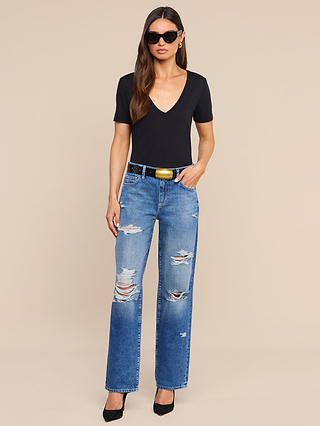 The woman is wearing the Brianna Cotton V-Neck Tee in black, paired with ripped blue jeans, a black belt with a gold buckle, black heels, and sunglasses as she stands against a plain beige background.