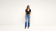 Woman wearing sunglasses, a black V-neck top, ripped blue jeans, and black heels stands against a plain white background with one hand in her pocket.
