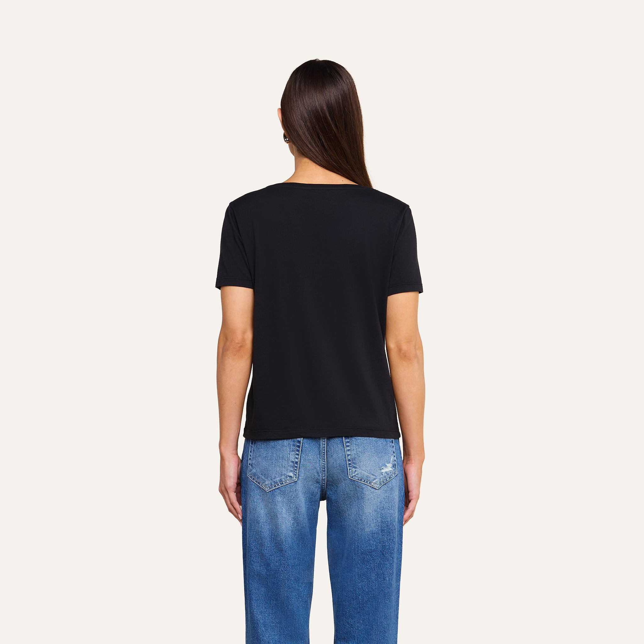 A person with long brown hair wears a black Brianna Cotton V-Neck Tee and blue jeans, standing with their back to the camera against a plain beige background.