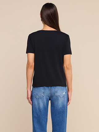 A person with long brown hair wears a black Brianna Cotton V-Neck Tee and blue jeans, standing with their back to the camera against a plain beige background.