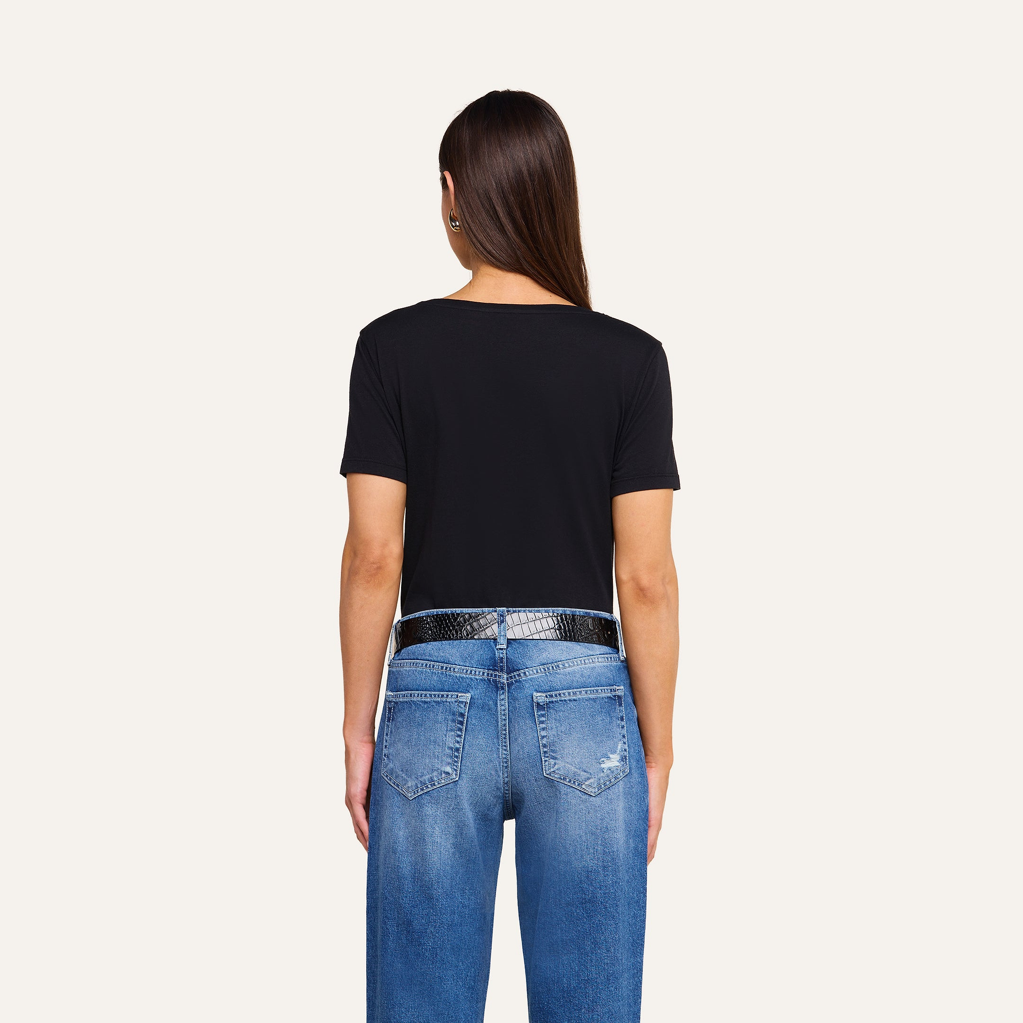 A person with long brown hair wears a black Brianna Cotton V-Neck Tee made from Supima cotton, styled with a black textured belt and blue jeans featuring visible back pockets and slight distressing, standing against a beige background and facing away from the camera.