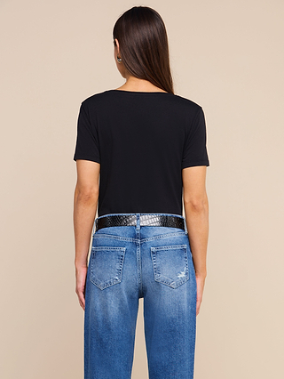 A person with long brown hair wears a black Brianna Cotton V-Neck Tee made from Supima cotton, styled with a black textured belt and blue jeans featuring visible back pockets and slight distressing, standing against a beige background and facing away from the camera.
