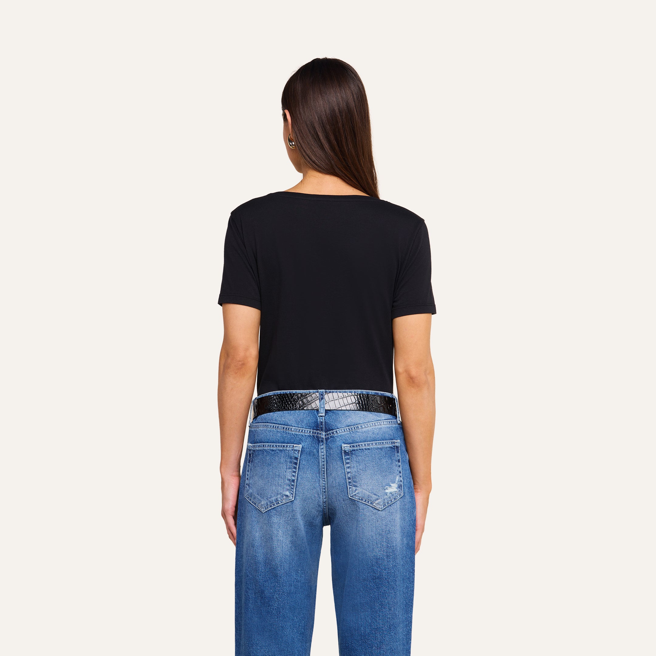 A person with long brown hair wears a black Brianna Cotton V-Neck Tee made from Supima cotton, styled with a black textured belt and blue jeans featuring visible back pockets and slight distressing, standing against a beige background and facing away from the camera.