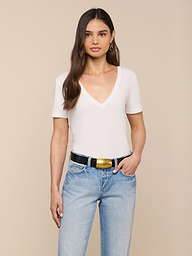 A woman with long brown hair wears the Brianna Cotton V-Neck Tee in white, light blue jeans, a black belt with a gold buckle, and gold hoop earrings while standing against a beige background.