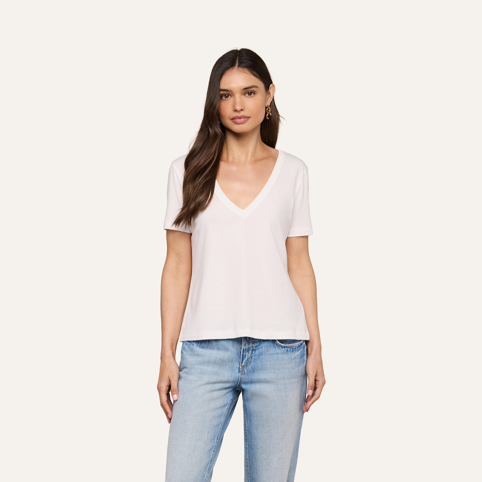 A woman with long brown hair wears the Brianna Cotton V-Neck Tee in white and light blue jeans, standing against a plain beige background and looking at the camera with a neutral expression.