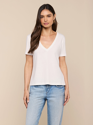 A woman with long brown hair wears the Brianna Cotton V-Neck Tee in white and light blue jeans, standing against a plain beige background and looking at the camera with a neutral expression.