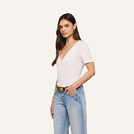 A woman with long brown hair stands against a beige background wearing the Brianna Cotton V-Neck Tee, light blue jeans, a black belt with a gold buckle, and gold hoop earrings. She poses with one hand in her pocket.