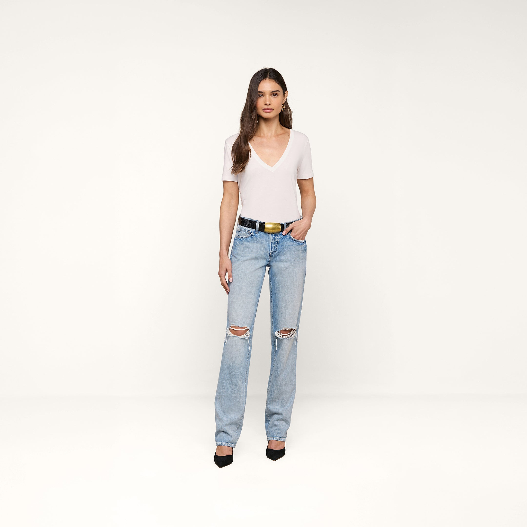 A woman with long brown hair stands against a beige background wearing the Brianna Cotton V-Neck Tee in white, light blue ripped jeans, a black belt with a gold buckle, and black heels. She has a relaxed expression and one hand in her pocket.