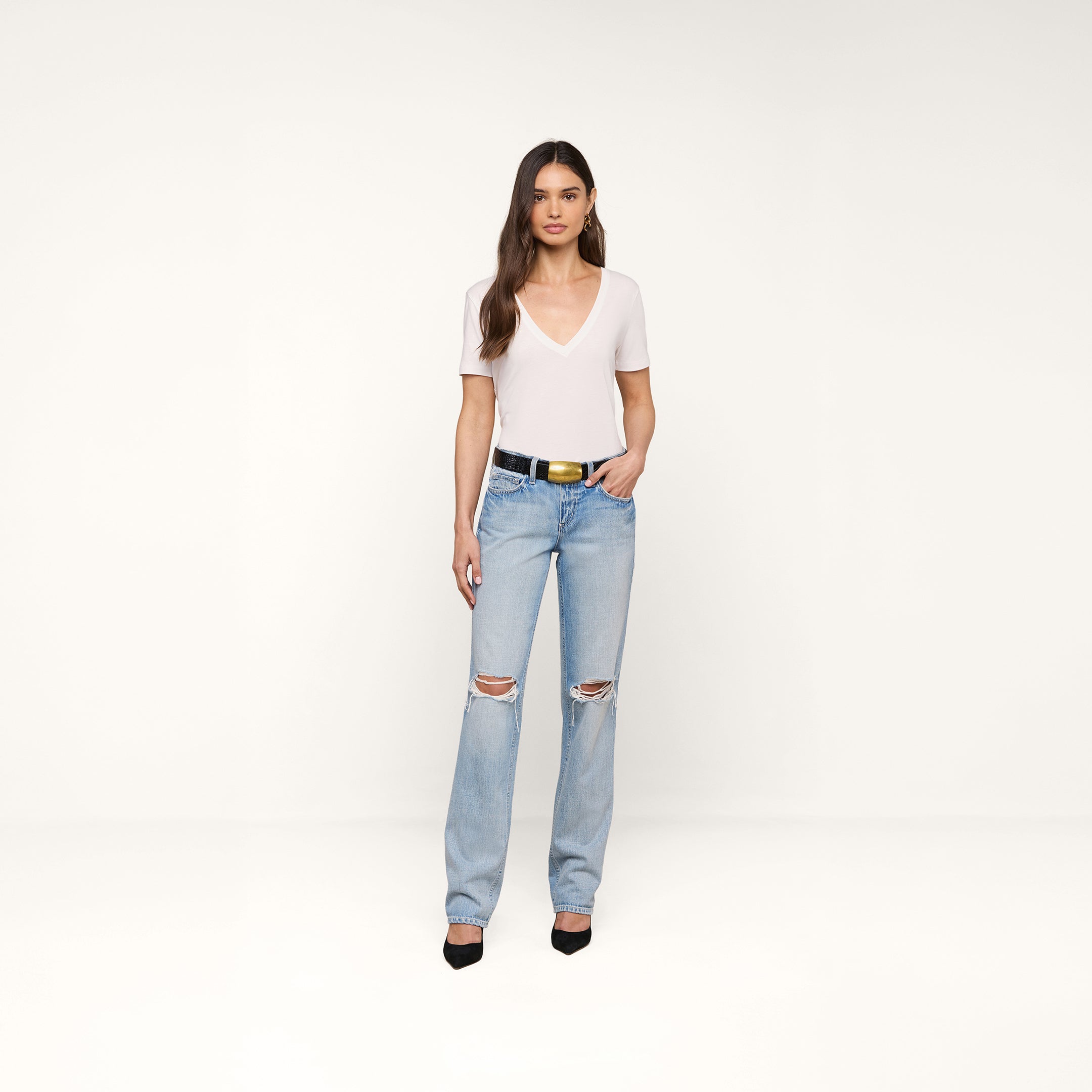 A woman with long brown hair stands against a beige background wearing the Brianna Cotton V-Neck Tee in white, light blue ripped jeans, a black belt with a gold buckle, and black heels. She has a relaxed expression and one hand in her pocket.