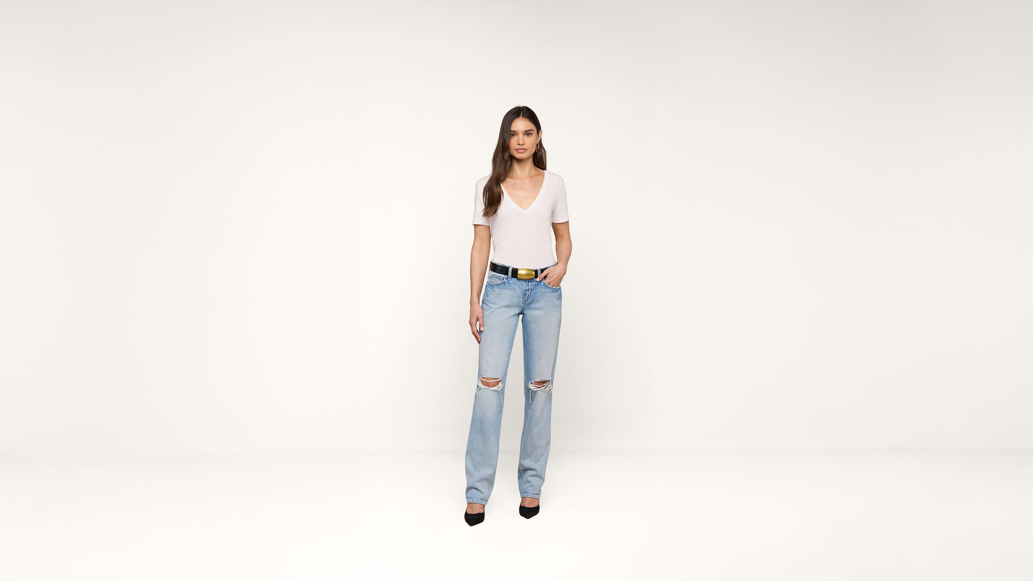Woman wearing a white V-neck T-shirt, ripped light blue jeans, and black heels stands against a plain white background with one hand in her pocket.