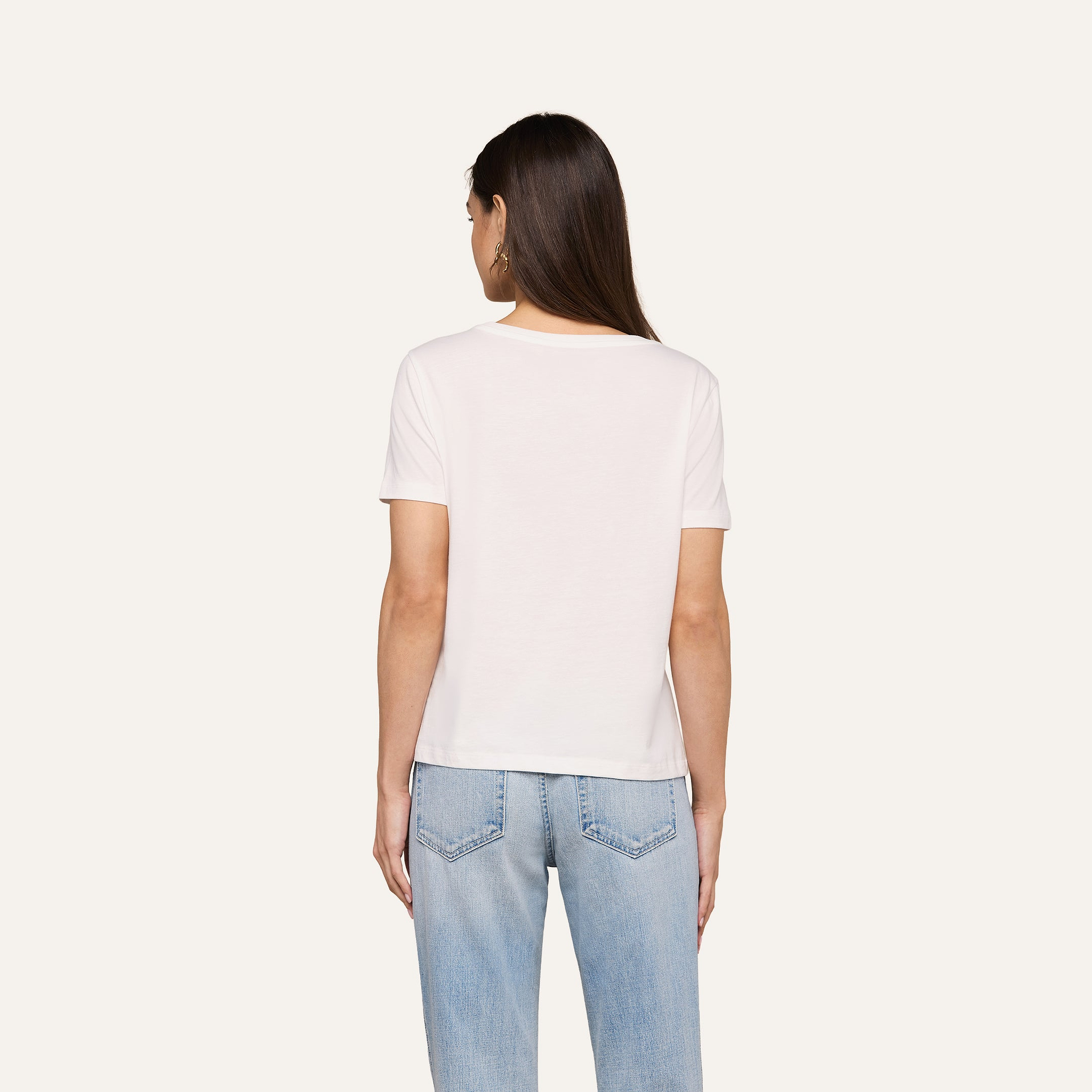 A woman with long brown hair stands with her back to the camera, wearing a relaxed fit white Brianna Cotton V-Neck Tee and light blue jeans against a beige background.