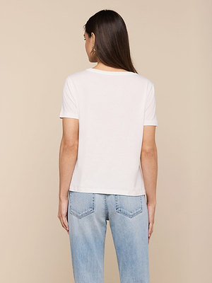 A woman with long brown hair stands with her back to the camera, wearing a relaxed fit white Brianna Cotton V-Neck Tee and light blue jeans against a beige background.