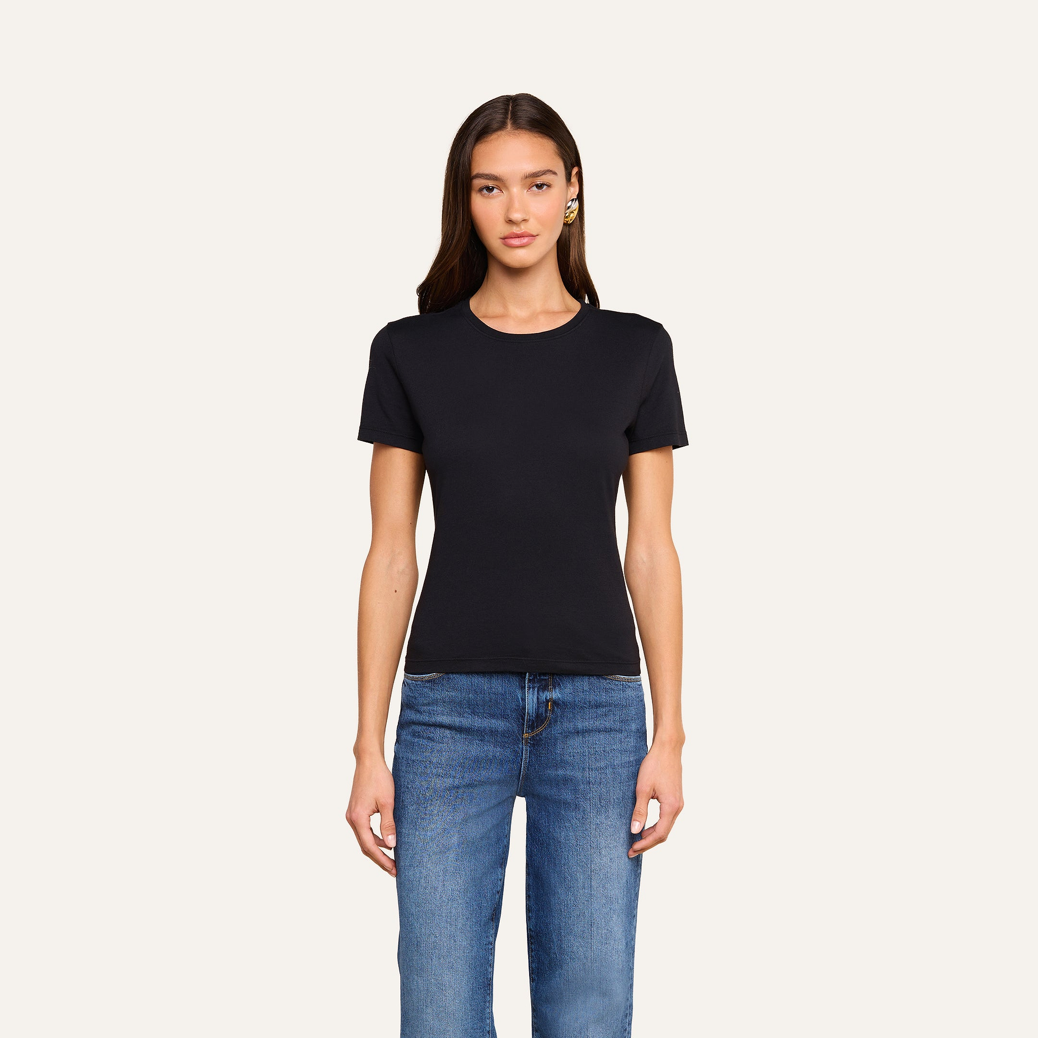 A woman with long brown hair stands facing forward against a beige background, wearing the Aileen Cotton Crewneck Tee in black with a relaxed fit and blue jeans.