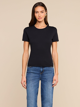 A woman with long brown hair stands facing forward against a beige background, wearing the Aileen Cotton Crewneck Tee in black with a relaxed fit and blue jeans.