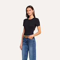 A woman with long brown hair wears the Aileen Cotton Crewneck Tee in black—a relaxed fit crewneck made from Supima cotton—paired with blue jeans, a brown belt, and gold earrings. She stands against a plain beige background with one hand in her pocket.